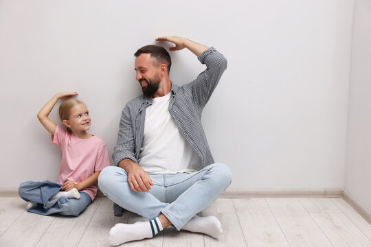 Father and daughter measuring their heights near light wall indoors. Space for text - Powered by Adobe
