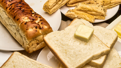 A close-up, high-quality image of freshly baked golden-brown bread loaf, soft white bread slices topped with a cube of butter, and crisp puff pastries in the background.