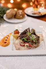 Tender roasted meat medallions on creamy sauce with peppers and herbs, elegant winter dinner plate, blurred golden holiday lights in background.