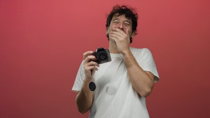Hispanic man laughing with a camera in hand against a bold red background, capturing a lively moment of humor and creativity.