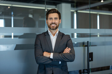 Businessman with a beard and dark suit standing confidently with arms crossed in a modern office...