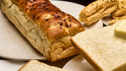 Close-up image of a freshly baked golden brown bread loaf with soft white bread slices topped with a cube of butter. Crisp puff pastries are visible in the background, creating a warm and inviting