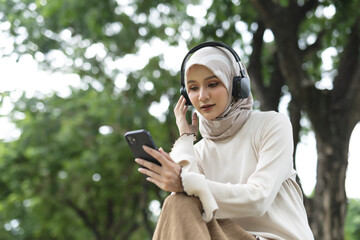 Muslim women in hijab wearing headphones, enjoying music or podcast on smartphone outdoors....