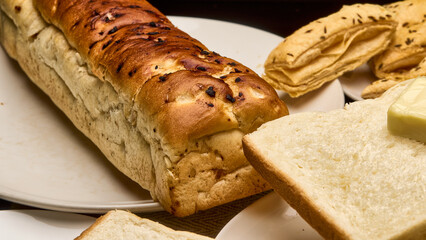 A close-up, high-resolution image of a freshly baked golden-brown bread loaf placed beside soft white bread slices topped with a cube of butter. Crisp puff pastries are visible in the background