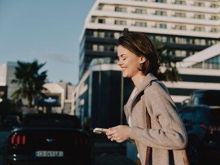 A cheerful business professional walks outdoors, texting on a smartphone. She smiles warmly,...