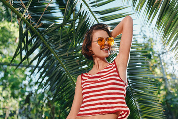 A cheerful woman wearing a striped crop top and sunglasses stands in a sunny tropical garden,...