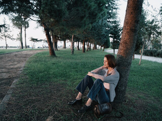 A contemplative woman sits beside a tree in a tranquil park. Leaning against the trunk, she wears...