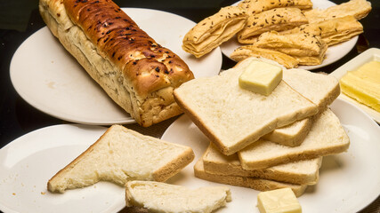 A close-up image showcasing a freshly baked golden-brown bread loaf, soft white bread slices stacked with a cube of butter on top, and crisp puff pastries in the background.