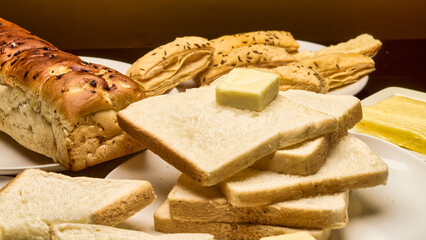 A close-up image of freshly baked golden-brown bread loaf, soft white bread slices topped with a cube of butter, and crispy puff pastries in the background with a warm lighting and detailed texture