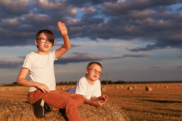 Two boys relaxing on hay bale in harvested field under dramatic sky. Concept of childhood friendship, simplicity, and nature connection.