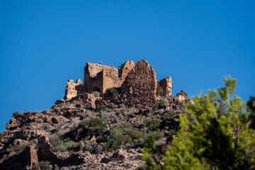 Remains of a historic stone fortress situated on a rocky hilltop surrounded by Mediterranean vegetation, set against a vibrant blue sky. This evocative scene captures the timeless character of rural