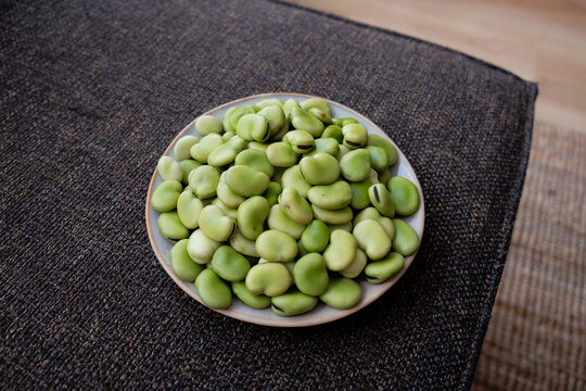 High resolution image of a ceramic bowl filled with fresh green fava beans placed on a textured dark fabric surface. Ideal for use in healthy eating, plant-based nutrition, seasonal vegetables, or