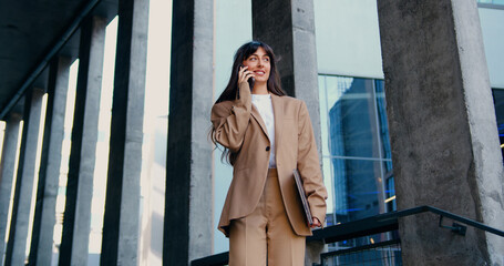 Fototapeta premium Confident female professional standing outside a modern office building, holding a laptop in one hand and speaking on the phone with a colleague, engaged in a serious business conversation