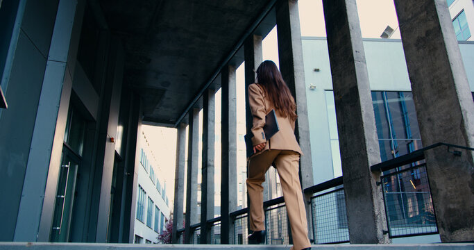Rear view of confident businesswoman with long flowing hair, wearing tailored formal suit, walking up outdoor stairs while holding laptop in one hand, approaching modern office building