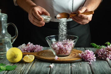 A chef adds sugar to a bowl filled with edible flowers, surrounded by fresh lemon and herbs. The warm lighting sets a cozy atmosphere in the rustic kitchen