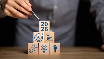 Stacked wooden blocks with icons of target, gear, chart, idea, and human symbol, topped with 2026 block, represent business vision, goal setting, innovation, productivity, and leadership planning.
