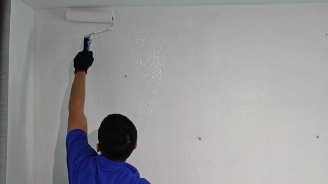 A young man paints a wall with a roller indoors.