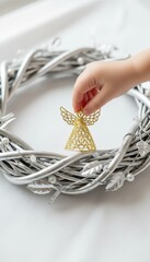 Fototapeta premium A child's hand placing a golden angel ornament on a silver Christmas wreath. Festive holiday decorating and family traditions. Minimalist background with copy space for text