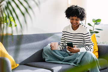 Young happy woman using mobile phone while enjoying hot tea or coffee drink relaxing on the sofa in...