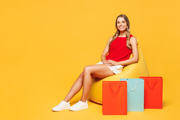Full body young smiling woman wearing red casual clothes hold in hand paper package bags after shopping sit in bag chair isolated on plain orange yellow background. Black Friday sale buy day concept.