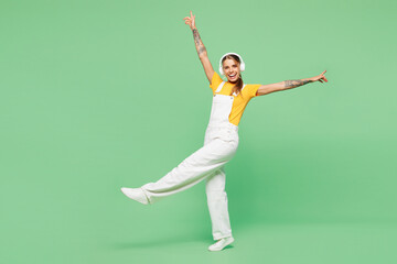 Fototapeta premium Full body young happy woman she wear yellow t-shirt overall casual clothes listen to music in headphones raise up hands dance isolated on plain pastel light green background studio. Lifestyle concept.