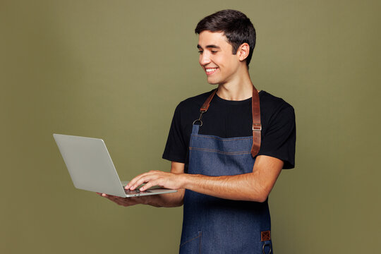 Young happy IT man barista bartender barman employee wear jeans apron black t-shirt work in coffee shop hold use laptop pc computer isolated on plain green background. Small business startup concept.