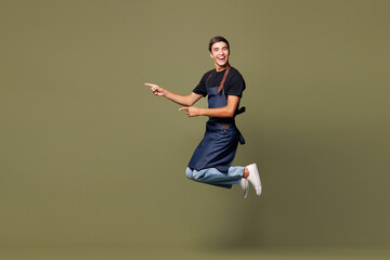 Full body young man barista bartender barman employee wear jeans apron t-shirt work in coffee shop jump high point index finger aside isolated on plain green background Small business startup concept © ViDi Studio