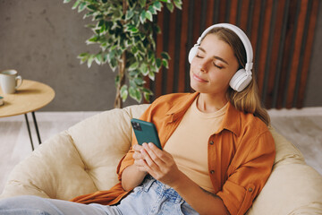 Young woman wear casual clothes listen music in headphones use mobile cell phone close eyes sit in chair stay at home hotel flat rest relax spend free time in living room indoor People lounge concept