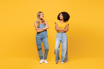 Full body two young friends buddies women wearing denim casual clothes together stand look to each other talk speak smile isolated on plain yellow orange background studio portrait. Lifestyle concept.