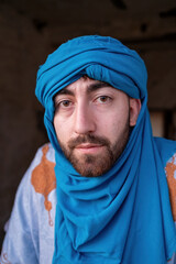 A close-up portrait of a Berber man wearing a vivid blue turban and traditional robe, looking directly at the camera inside an old rustic house.