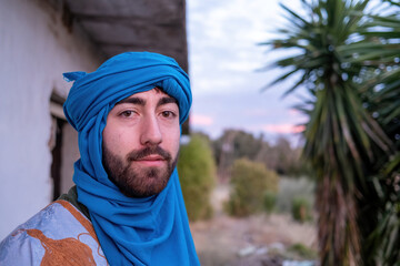 A Berber man dressed in a blue turban and traditional embroidered robe stands outdoors near a...