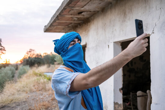 A Berber man wearing a bright blue turban and traditional robe takes a selfie with his smartphone beside an old rural house at sunset. - Powered by Adobe