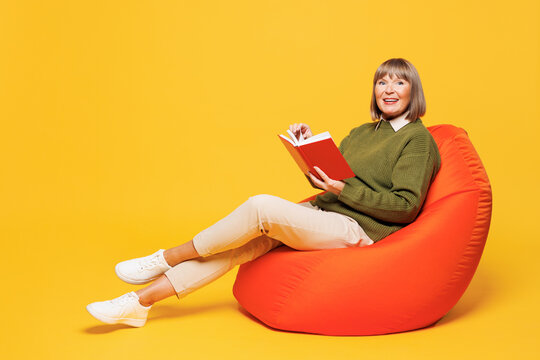 Full body side view elderly smart blonde woman she wears green sweater casual clothes sit in bag chair reading novel book isolated on plain yellow orange background studio portrait. Lifestyle concept.