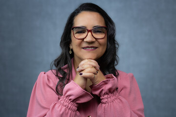 Adult woman with long gray hair, wearing eyeglasses and pink blouse, smiling with hands in prayer position. Horizontal studio portrait with copy space, perfect for religion, faith, belief, gratitude.