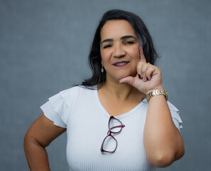 Mature adult woman with hand touching face, wearing white shirt, eyeglasses hanging on her neckline. Studio portrait with copy space, suitable for women's issues, femininity, age, or facial skincare.