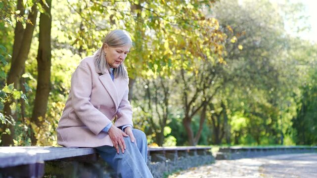 Elderly female with gray hair suffers from painful severe knee joint pain sitting on bench on street in urban city park. Upset woman massages leg muscles, could not walk, illness, cramps or rheumatism