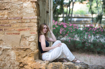 A young, pretty blonde woman wearing white trousers and a black cotton shirt is sitting on a stone wall of a ruined building in the forest. The woman is resting and touching her head with her hand.