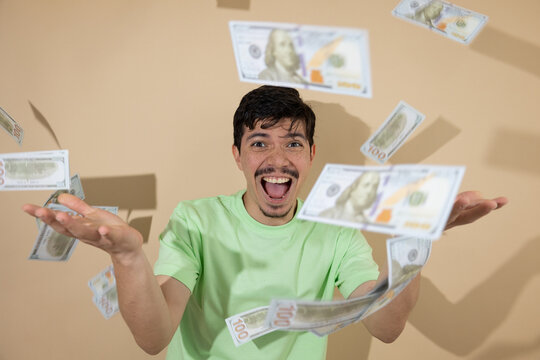 Smiling freckled young man in a green shirt extending his hands as bills fall against a light brown background. Ideal image for concepts of financial success and economic prosperity