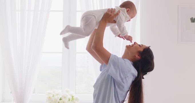 Happy mothers hands lift baby up, smiling mom lifting newborn child into air, playing, kissing with love and care. Young laughing woman standing at home window to carry little toddler and hug