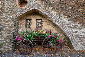A rustic wooden cart filled with bright pink and colorful flowers is displayed under a stone archway of the historic Reichsburg Cochem castle wall in Germany.