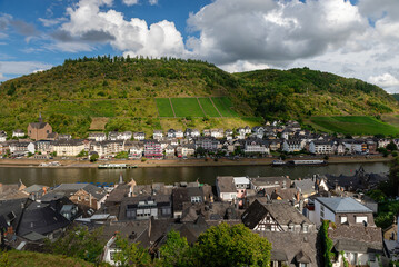 Elevated panoramic view of the town of Cochem along the Moselle River, featuring historic rooftops in the foreground, and steep terraced vineyards on the surrounding hills under a bright, cloudy sky