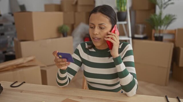 Woman using phone and credit card indoors surrounded by moving boxes suggests recent relocation to a new home or apartment interior while communicating and multitasking.
