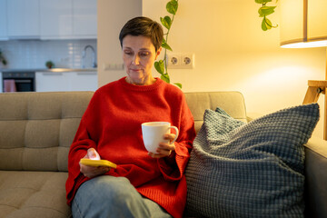 Contemplative older woman browsing social media, drinking hot tea on cozy sofa. Evening home rest,...