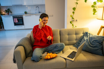 Cheerful female multitasks with snack screen and call in cozy home. Happy mature woman watching tv,...