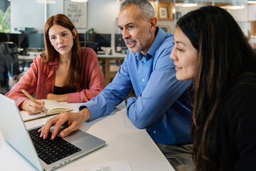 Mid adult businessman working together with two young female colleagues on a laptop in a modern...