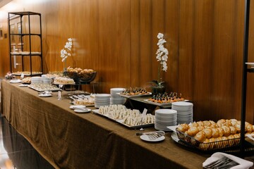 Long buffet table laden with diverse small sandwiches, savory appetizers, pastries, and stacks of clean white plates ready for guests in a conference hall