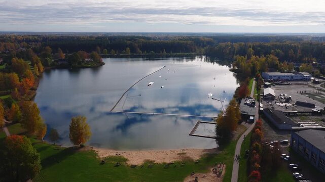 Aerial sweep view over a lakeside wakepark park in Jelgava, Latvia. Sandy beach, promenade, docks, floating barriers, wakeboarding obstacles, and autumn trees are visible.