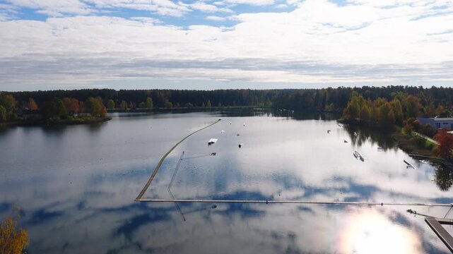 High angle aerial shows a mirror like lake, sandy beach, grassy park, cable system, ramps, docks, and buoys in Jelgava, Latvia, under soft midday light.