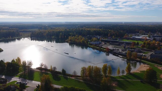 Aerial view over a calm lake in Jelgava, Latvia. Tree lined promenade, sandy beach, and wakepark obstacles with cable towers. Morning light casts long shadows.