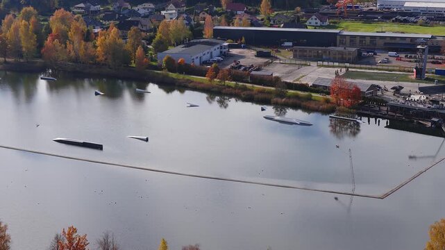 Aerial high angle view shows Jelgava lake with wakepark, Latvia, with cable wakeboarding, ramps and floating barrier as a rider moves, fall trees and buildings reflect in daylight